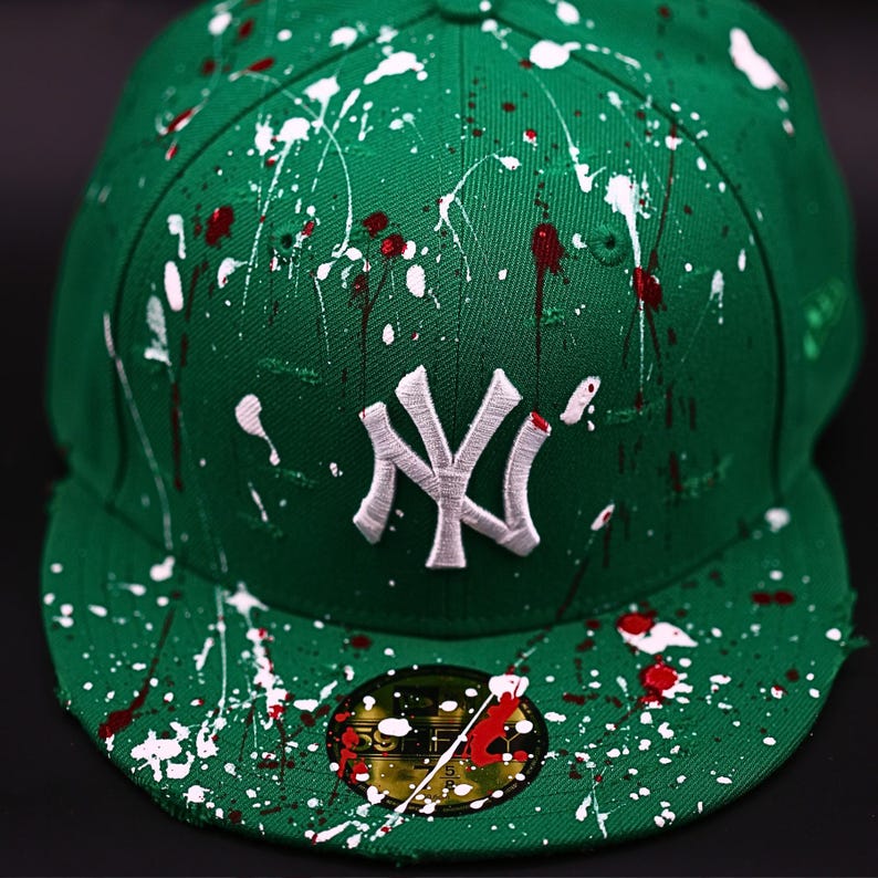 May include: Green baseball cap with white and red paint splatter design. Features a white embroidered "NY" logo on the front. The cap has a flat brim and a fitted design, with a small sticker on the brim.
