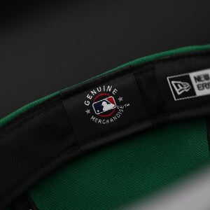 May include: A green baseball cap with a black brim, showing the inner label. The label reads "Genuine Merchandise" with the MLB logo. The cap also has a "59FIFTY" label and the New Era logo.