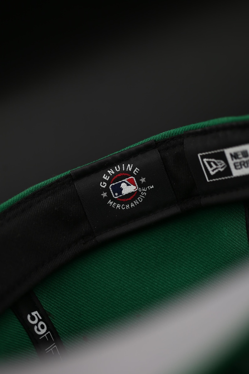 May include: A green baseball cap with a black brim, showing the inner label. The label reads "Genuine Merchandise" with the MLB logo. The cap also has a "59FIFTY" label and the New Era logo.