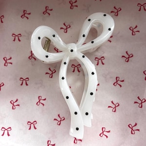 May include: A white bow-shaped hair clip with black polka dots. The hair accessory has a gold-coloured spring and is set against a pink background with red bow patterns. This hair clip is a stylish accessory.