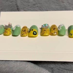 May include: Set of ten green and yellow press-on nails with glitter and a paw print design. One nail has a clear rhinestone.