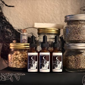 May include: Three amber glass dropper bottles with labels reading Mystical and Divine Sacred Earth featuring a crescent moon and cat illustration, alongside two glass jars filled with dried herbs, a small vial, and a larger jar with a gold lid.
