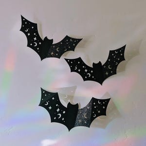 Puede incluir: Tres decoraciones de murciélagos negros con detalles de purpurina, estrellas y lunas crecientes se exhiben en una pared blanca. Estos murciélagos con temática de Halloween probablemente se utilizan para la decoración de temporada.