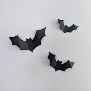 May include: Three black paper bats with geometric designs against a white backdrop. The bats vary in size, with detailed wing structures, ideal for Halloween or themed decorations.