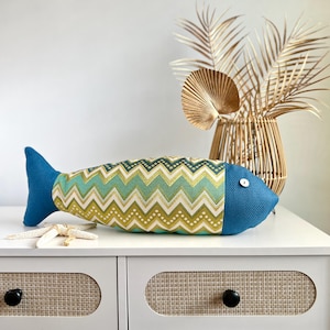May include: A blue and green chevron patterned fish-shaped pillow with a blue tail fin and a white button eye. The pillow is sitting on a white dresser with two drawers.