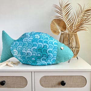 May include: A turquoise blue fish-shaped pillow with a white and blue wave pattern. The pillow is sitting on a white dresser with two drawers.