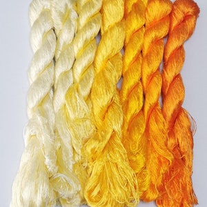 May include: A collection of silk threads in a gradient of yellow and orange hues. The skeins of thread are arranged side by side, transitioning from a pale cream color to vibrant shades of yellow and orange. The threads are tightly twisted, showcasing their texture.