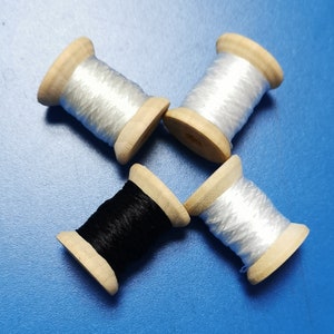 Könnte beinhalten: Vier Holzspulen mit Faden, drei mit weißem Faden und eine mit schwarzem Faden, sind auf einer blauen Oberfläche angeordnet. Die Spulen haben eine natürliche Holzoberfläche. Der Faden ist fest um die Spule gewickelt.