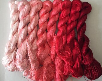 May include: A collection of silk thread skeins in a gradient of pink and red hues. The skeins transition from light pink to deep red, showcasing a range of shades. The threads are tightly wound, suggesting their use in embroidery or other textile crafts.