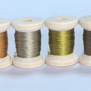 Puede incluir: Seis carretes de hilo de madera en fila, cada uno con un color diferente. Los colores incluyen gris, dorado, verde oliva y marrón. Los carretes son de madera clara.