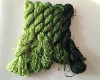 May include: Five skeins of green embroidery floss in varying shades, from light lime to deep forest green, are arranged side by side. The floss is tightly wound, with loose strands visible. The background is plain white.