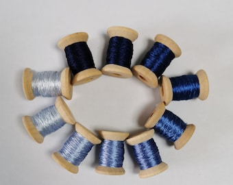 May include: A collection of wooden spools, each wound with a different shade of blue thread. The spools are arranged in a circular pattern, showcasing a range of blues from light silver to deep navy. The wooden spools add a rustic touch to the display.