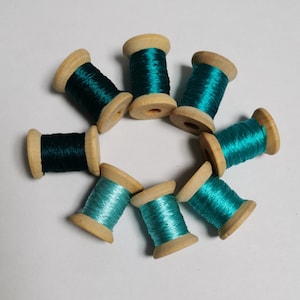 Puede incluir: Ocho carretes de madera llenos de hilo verde azulado y verde azulado oscuro, dispuestos en círculo. El hilo está enrollado alrededor de los carretes de madera, creando una exhibición visualmente atractiva. Ideal para coser y manualidades.