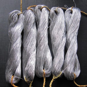 Può includere: Cinque matasse di filo metallico color argento, ciascuna legata con uno spago color naturale. Il filo è avvolto strettamente, creando un aspetto strutturato. Le matasse sono disposte in fila su uno sfondo scuro.