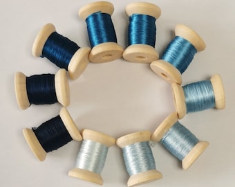 May include: A collection of ten wooden spools of thread in various shades of blue and silver. The spools are arranged in a circular pattern, with the thread wound around the wooden core. The image is taken from a top-down perspective.