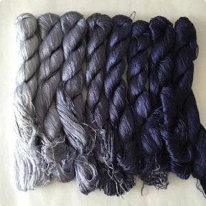 May include: A collection of silk yarn skeins in a gradient of gray and blue hues. The yarn is tightly twisted, with a soft sheen, transitioning from light silver to deep navy. The skeins are arranged side by side, showcasing the color variations.
