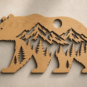 Könnte beinhalten: Hellbraune Wanddekoration in Bärenform aus Holz. Das Design zeigt ausgeschnittene Berge, Bäume und ein kreisförmiges Loch. Das Kunstwerk stellt einen Bären mit einer Berg- und Waldszene dar.