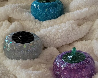 May include: Three small, glittery pumpkins in different colors: teal, white with iridescent glitter, and purple. The pumpkins are sitting on a white fluffy surface.
