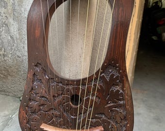 May include: A wooden lyre with a carved floral design. The lyre has 10 strings and a wooden bridge. The lyre is brown and has a natural finish.