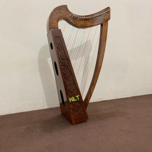 以下が含まれることがあります： 精巧な彫刻と弦を備えた木製のハープ。ハープは、フレームとサウンドボックスに沿って装飾的な模様が施された濃い茶色の仕上げです。弦は白く、ハープは茶色の表面に立っています。「KILT COUTURE」というテキストが表示されます。