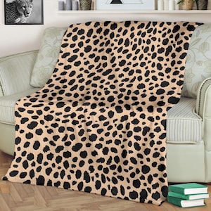 May include: A tan and black cheetah print blanket draped over a sofa. The blanket features a repeating pattern of black spots on a tan background. The sofa is cream-colored with floral patterned pillows.