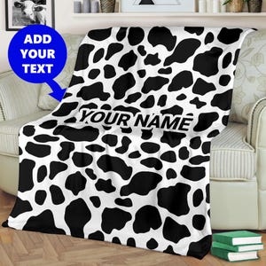 May include: Black and white cow print throw blanket with the text "YOUR NAME" printed on it.