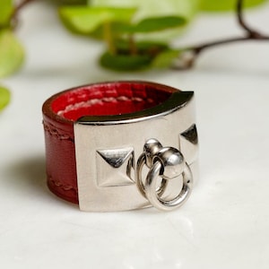 May include: A red leather ring with a silver metal band and a silver ring attached. The metal band has a series of pyramid-shaped studs.