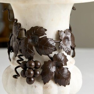 May include: A white marble pedestal with a decorative bronze grape vine design. The bronze vine features detailed leaves and a cluster of grapes. The pedestal has a rounded base and a wide top.