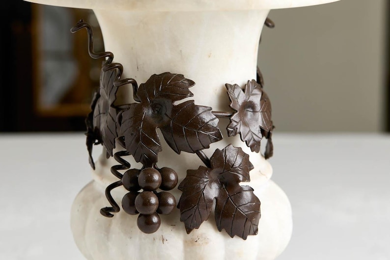 May include: A white marble pedestal with a decorative bronze grape vine design. The bronze vine features detailed leaves and a cluster of grapes. The pedestal has a rounded base and a wide top.