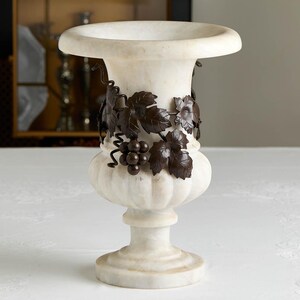May include: A white marble urn with a wide rim and a pedestal base. The urn is adorned with a dark bronze-colored grape vine design around the body. The urn is a decorative piece.