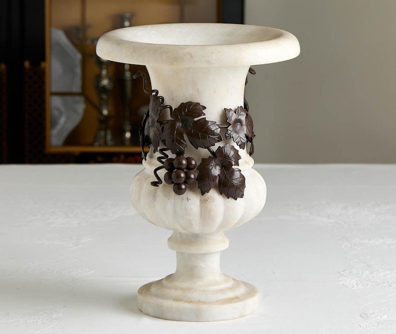 May include: A white marble urn with a wide rim and a pedestal base. The urn is adorned with a dark bronze-colored grape vine design around the body. The urn is a decorative piece.