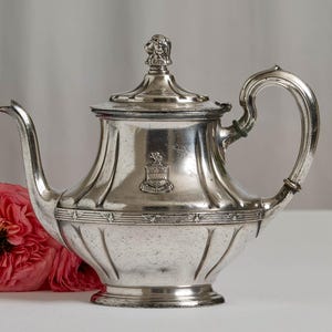 May include: A silver-toned metal teapot featuring a curved handle, a long spout, and a lid topped with a sculpted dog head finial. The body is decorated with vertical fluting and a central crest emblem featuring a lion and the text LIBERTAS.