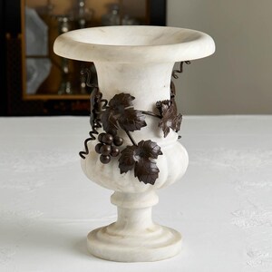 May include: A white marble urn with a wide rim and pedestal base. Dark brown metal grape vine and leaf accents wrap around the urn's body. The urn is a decorative piece for home decor.