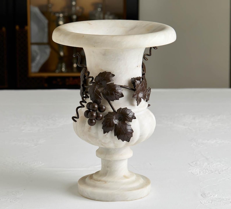 May include: A white marble urn with a wide rim and pedestal base. Dark brown metal grape vine and leaf accents wrap around the urn's body. The urn is a decorative piece for home decor.