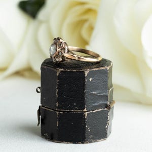May include: A vintage engagement ring with a rose cut diamond surrounded by smaller diamonds, set in a gold band. The ring is displayed in a black ring box.