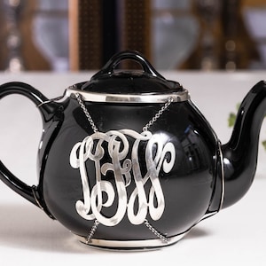 May include: Black teapot with a silver monogram on a chain. The teapot has a silver rim and a silver lid.