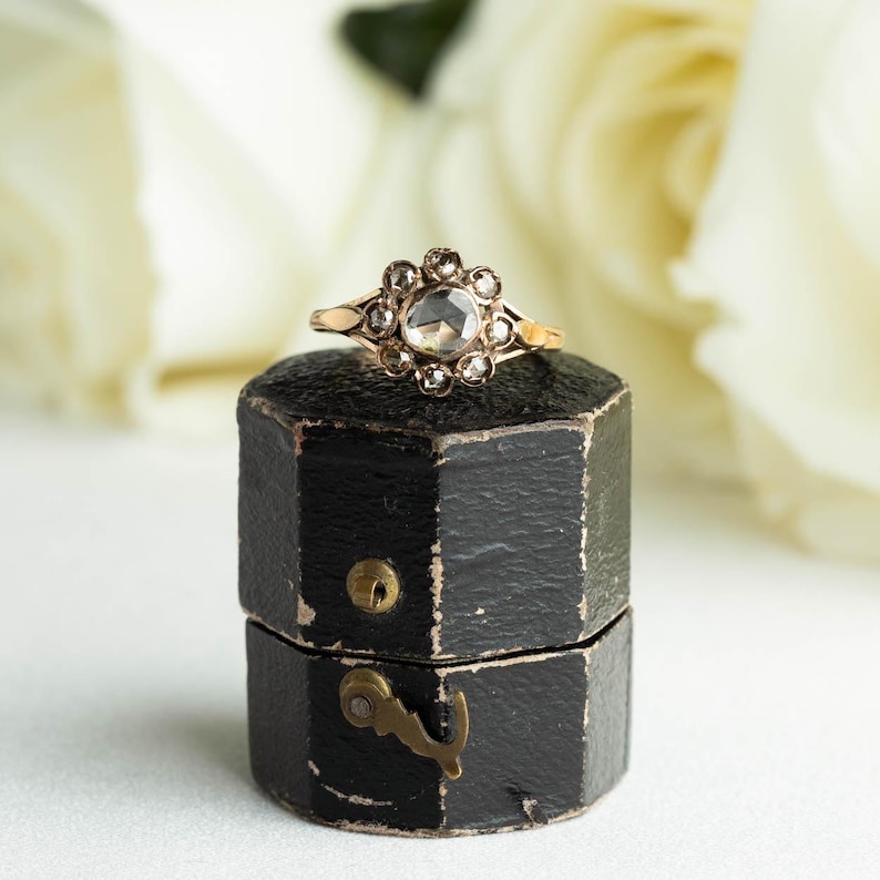 May include: A vintage ring with a round, clear stone surrounded by smaller diamonds, set in a gold band. The ring is displayed in a black, octagonal ring box.