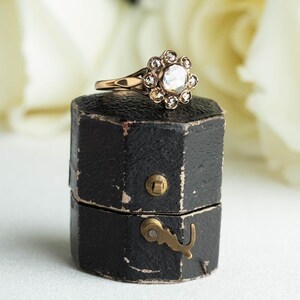 May include: A vintage ring with a large, round, clear stone surrounded by smaller stones in a floral design. The ring is in a black ring box with a brass latch.