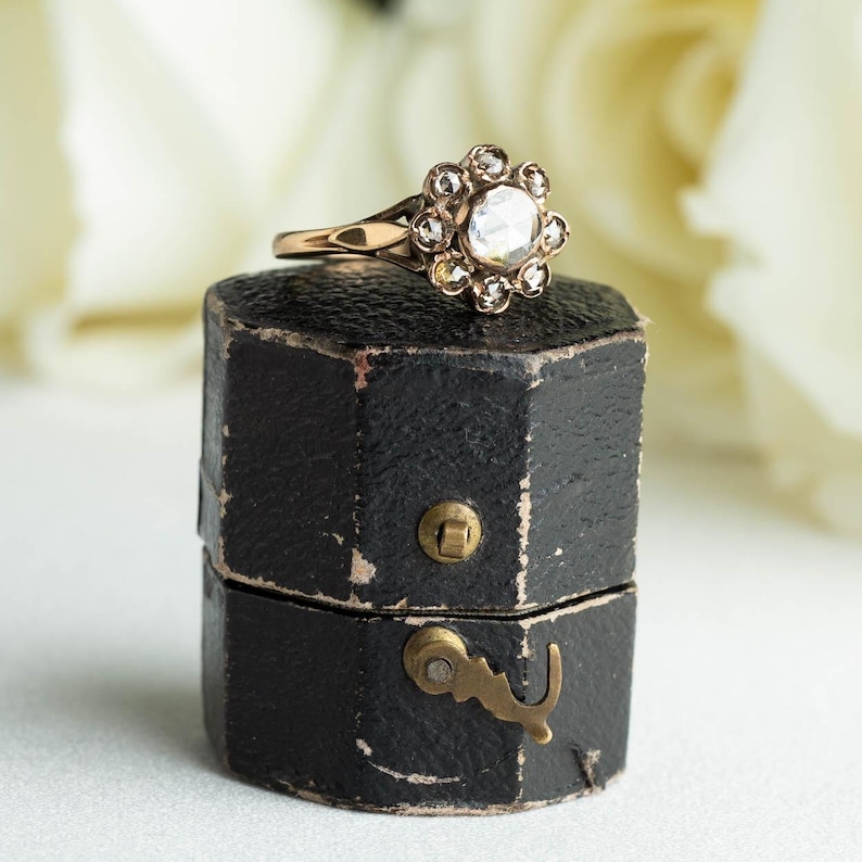 May include: A vintage ring with a large, round, clear stone surrounded by smaller stones in a floral design. The ring is in a black ring box with a brass latch.
