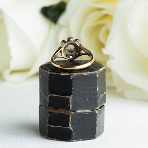 May include: A vintage ring with a round, clear stone surrounded by smaller stones. The ring is set in gold and is sitting in a black ring box.