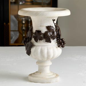 May include: A white marble vase with a wide rim and a pedestal base. The vase is adorned with bronze-colored grape vines and clusters of grapes. The vase is a decorative item.