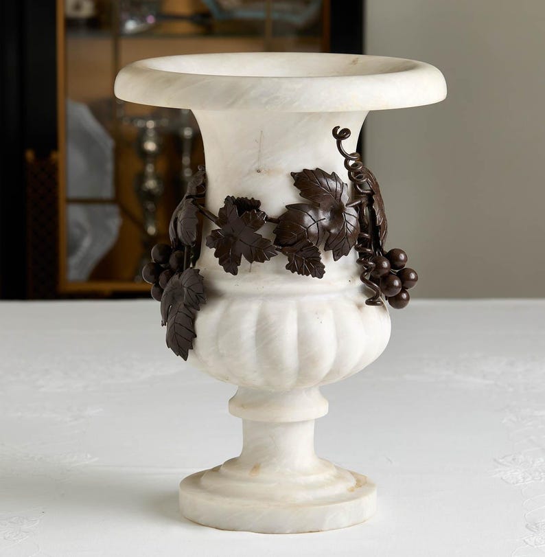 May include: A white marble vase with a wide rim and a pedestal base. The vase is adorned with bronze-colored grape vines and clusters of grapes. The vase is a decorative item.