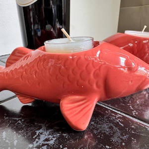 May include: Two red ceramic fish-shaped candle holders. Each holder has a cavity for a small tea light candle. The fish have detailed scales and fins. A dark bottle is in the background.