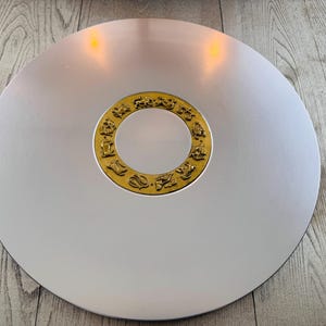 May include: A large, silver-colored decorative plate with a gold-colored ring in the center. The ring features zodiac symbols. The plate is round and appears to be made of metal. The plate is on a wooden surface.