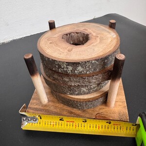 May include: A set of four wooden coasters made from tree slices, stacked in a holder. The coasters have a natural bark edge and a hole in the center. The holder has four wooden dowels. A yellow tape measure is in the foreground.
