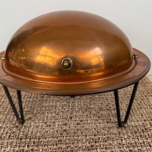 May include: A copper-colored serving dish with a domed lid, resting on a black metal stand. The dish has a circular base and a shiny, reflective surface. The stand has three angled legs.