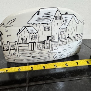 May include: A white ceramic decorative piece with a black and white illustration of seaside houses, a fence, and a flying bird. The piece is approximately 8 inches long.