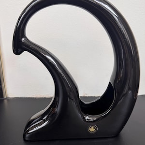 May include: A glossy black ceramic wine bottle holder with a unique, curved design. The holder features a large, open loop and a base with a small, circular emblem. The overall shape is modern and sculptural.
