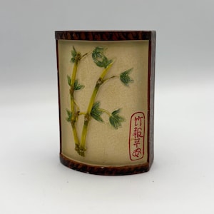 May include: A semi-cylindrical decorative container with a cream-colored front panel featuring a raised relief of green and yellow bamboo stalks. The frame is dark brown with a textured finish. A red rectangular stamp contains the text 竹報平安.