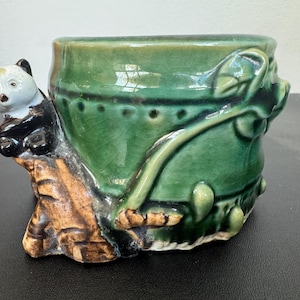 May include: A ceramic planter with a green glazed finish. A panda bear figure is perched on a tree trunk on the side of the planter. The planter has a textured surface and is decorated with leaf and vine details.